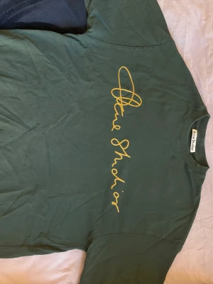 Grön Acne Studios t-shirt med tryck - Snygg grön t-shirt från Acne Studios med gul text i ett handskrivet typsnitt över bröstet. Klassisk rund halsringning och korta ärmar. Perfekt för dig som gillar stilrena plagg med en twist. Materialet är mjuk bomull.