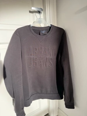 Svart sweatshirt från Armani Jeans - Svart sweatshirt med broderad logga och texten 'Armani Jeans' framtill. Tröjan har rund halsringning och långa ärmar. Perfekt för en stilren och avslappnad look.