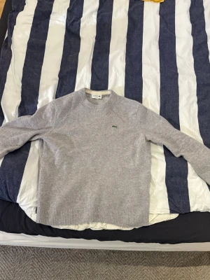 Grå stickad tröja från Lacoste - Snygg grå stickad tröja från Lacoste med rund halsringning och det klassiska gröna krokodilmärket broderat på bröstet. Tröjan har ribbade muddar vid ärmslut och nederkant. Perfekt för lager på lager och enkel att matcha.