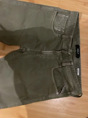 Olivgröna chinos från Replay 34/36 - Snygga olivgröna chinos från Replay med smal passform och klassisk femficksdesign. Byxorna har bälteshällor och är tillverkade i ett mjukt bomullsmaterial som ger en skön känsla. Perfekta för dig som gillar stilrena och bekväma byxor.
