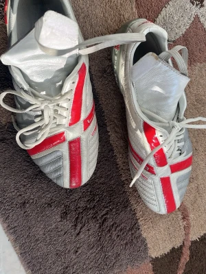 Umbro X-Boot rödvita fotbollsskor - Säljer ett par Umbro X-Boot fotbollsskor i storlek 42.5. Skorna är vita med röda detaljer i ett korsmönster och har klassisk snörning. Ovandelen är i syntetmaterial med läderkänsla och skon har en låg profil som passar för snabba rörelser på planen.