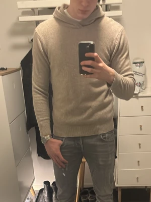 Beige stickad hoodie i merinoull - Snygg beige stickad hoodie med lång ärm och ribbade muddar. Tröjan har en avslappnad passform och är tillverkad i mjuk merinoull, perfekt för lager-på-lager. Enkel och stilren design utan synligt märke.