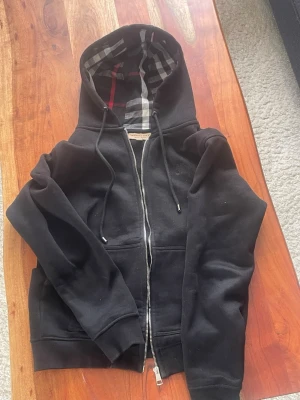 Svart hoodie från Burberry (dragkedjan har en stud borta) - Svart hoodie från Burberry med klassiskt rutigt foder i huvan och dragkedja framtill. Hoodien har dragsko vid huvan och två fickor framtill. Perfekt för dig som gillar stilrena och exklusiva plagg med en touch av brittisk design.