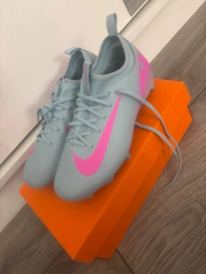 Nike Mercurial blå och rosa fotbollsskor - Snygga Nike Mercurial fotbollsskor i ljusblått med stora rosa swoosh-loggor och detaljer. Skorna har snörning, låg profil och är tillverkade i syntetmaterial för bra bollkontroll och lätt känsla. Perfekta för dig som vill sticka ut på planen. Helt nya.
