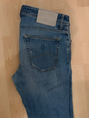 Ljusblå jeans från Jacob Cohën - Säljer ett par ljusblå jeans från Jacob Cohën med raka ben och klassisk femficksmodell. Jeansen har knäppning med knappar framtill och en ljus patch med logga bak i midjan. Tillverkade i mjukt denimtyg med diskreta slitningar för en avslappnad look.