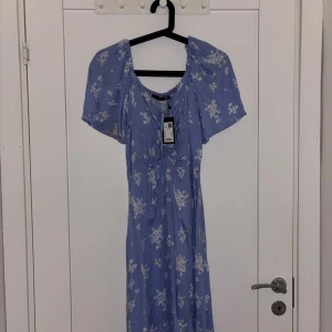Blå blommig långklänning  - Supersöt blå långklänning från lager med vitt blommigt mönster. Klänningen har korta ärmar, v-ringning och lätt volang vid fållen. Perfekt för sommarens alla tillfällen!