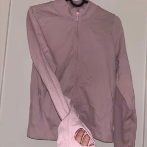 Ljusrosa zip-tröja med tumhål - Snygg ljusrosa träningsjacka med dragkedja framtill och tumhål i ärmarna. Jackan har en smal passform och hög krage, perfekt för löpning eller gym. Materialet känns stretchigt och mjukt mot huden.