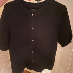 Svart kofta med kort ärm från Gina Tricot - Säljer en svart kofta från Gina Tricot med korta ärmar och rund hals. Koftan har knappar framtill och är stickad i mjukt material. Perfekt att styla över en topp eller klänning.