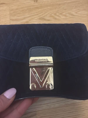 Svart axelremsväska från Valentino - Snygg svart handväska från Valentino i mjuk sammet med quiltat mönster. Väskan har en guldfärgad metallspänne med logotyp och kedjerem i metall. Perfekt för dig som vill ha en stilren och lyxig accessoar.