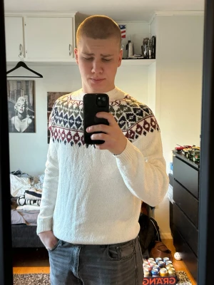 Vit stickad tröja med mönster - Säljer en vit stickad tröja med klassiskt nordiskt mönster i svart, vinrött och mörkgrönt runt axlarna. Tröjan har rund hals och ribbade muddar vid ärmslut och nederkant. Perfekt för kalla dagar och snygg att matcha med jeans.