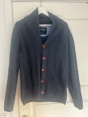 Mörkgrå kofta från Bondelid - Mysig mörkgrå kofta från Bondelid med grovstickad struktur, stora träknappar och bred krage. Broderad logga på bröstet och brittisk flagga på ärmen ger en cool detalj. Perfekt för lager på lager och chill stil.