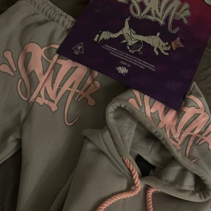 Grå hoodie från SYNA WORLD med rosa tryck - Säljer en grå hoodie från syna world med märket SYNA på luvan och byxan. Finns 1-2 små fläckar men syns inte.