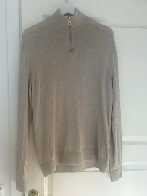 Beige halvzip tröja med hög krage - Stilren beige tröja med halv dragkedja och hög krage. Långärmad modell med ribbade muddar och nederkant. Perfekt för lager-på-lager och enkel att matcha med olika outfits. Endast provad då jag ej hann lämna tillbaka efter beställning.