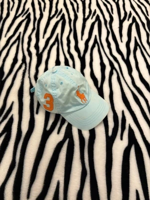 Polo Ralph Lauren Keps - Snygg ljusblå keps från Polo Ralph Lauren med orange broderad logga och en stor orange siffra 3 på sidan. Justerbar rem baktill. Perfekt accessoar för en sportig och avslappnad stil.