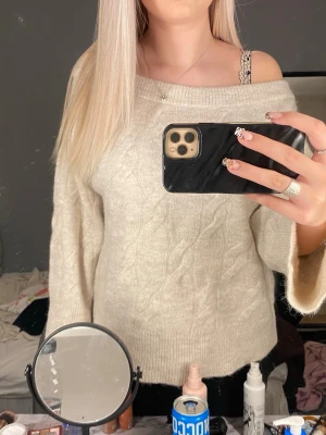 Beige stickad off shoulder-tröja - Snygg beige stickad tröja med kabelstickat mönster och off shoulder-design. Tröjan har långa ärmar och bred ribbad kant nedtill och vid ärmsluten. Perfekt för en chill och trendig look.