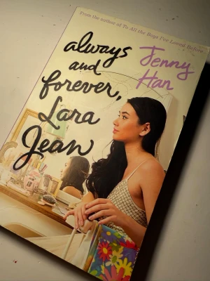 Always and Forever, Lara Jean av Jenny Han - Säljer boken 'Always and Forever, Lara Jean' av Jenny Han. Omslaget är ljust med svart och lila text, och visar en tjej vid ett skrivbord med blommigt mönster. Perfekt för dig som gillar romantisk ungdomslitteratur. På engelska