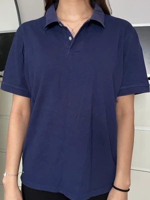 Marinblå pikétröja med krage - En enkel och stilren marinblå pikétröja med klassisk krage och korta ärmar. Tröjan har två knappar vid halsen och är gjord i mjukt bomullsmaterial. Perfekt för en avslappnad och snygg look.