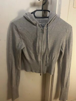 Sister Knitwear - Snygg grå croppad hoodie med dragkedja framtill och ribbade muddar. Hoodien har justerbar luva med snören och bred ribbad kant nedtill. Perfekt för en chill och trendig look. Materialet känns mjukt och stretchigt.