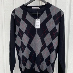 Argyle stickad tröja från Dressmann - Snygg stickad tröja från Dressmann med klassiskt argylemönster i svart, grått och tunna röda linjer. Tröjan har V-ringning hel svart baksida. Helt ny med lappar och från ett djur och rökfritt hem! 