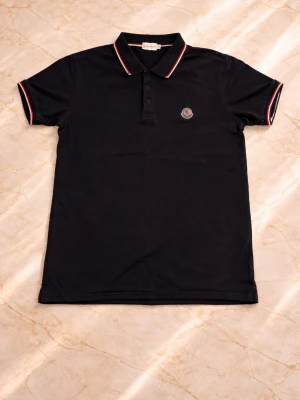 Mörkblå Moncler pikétröja med röd detalj - Klassisk svart pikétröja från Moncler med röd och vit rand på krage och ärmslut. Tröjan har korta ärmar, knappslå framtill och Moncler-logga på bröstet. Perfekt till jeans eller shorts för en clean och stilren look.