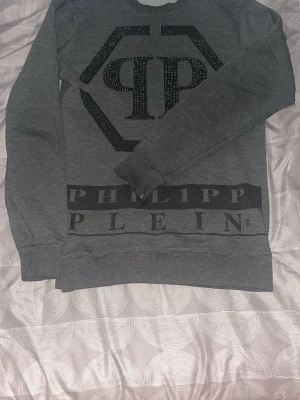 Grå sweatshirt från Philipp Plein - Snygg grå sweatshirt från Philipp Plein med stort svart logotryck och glittrande detaljer framtill. Tröjan har rund halsringning och långa ärmar. Perfekt för en trendig och avslappnad stil.