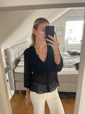 Svart transparent blus med volang - Snygg marinblå blus med volangdetaljer framtill och knappar. Blusen är transparent och har trekvartsärm. Perfekt att matcha med jeans eller kjol för en cool look. Materialet känns lätt och luftigt.