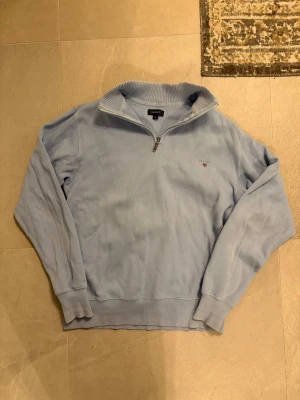 Ljusblå stickad tröja från GANT - Säljer en ljusblå stickad tröja från GANT med halv dragkedja och ribbad krage. Tröjan har broderad logga på bröstet och är tillverkad i mjukt material. Perfekt för lager på lager och chill stil.