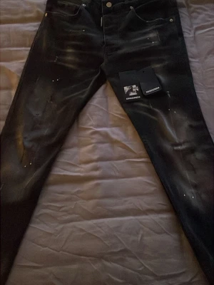 Svarta Dsquared2 jeans med slitningar - Säljer ett par svarta jeans från Dsquared2 med coola slitningar och färgstänk för en trendig look. Jeansen har klassisk femficksmodell och knappgylf. Perfekta för dig som vill sticka ut med en edgy stil.