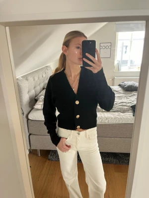 Svart kofta med stora knappar - Säljer en svart, kort kofta med v-ringning och stora, guldiga knappar framtill. Koftan har långa ärmar och ribbade muddar. Perfekt att slänga över en topp för en snygg look.