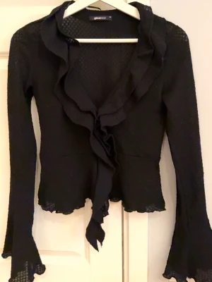 Svart volangblus från Gina Tricot - Supersnygg svart blus från Gina Tricot med volangkant längs ringningen och framtill. Blusen har lång ärm med utsvängda ärmslut och lätt transparent struktur. Figurnära passform och v-ringning ger en trendig look. Perfekt att styla till jeans eller kjol.