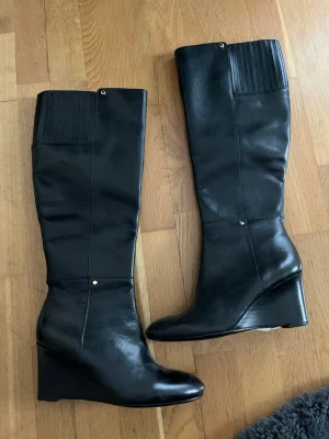 Svarta kilklackade stövlar  - Snygga svarta stövlar med kilklack och högt skaft. Tillverkade i slätt läder med elastiska detaljer upptill för bättre passform. Klassisk och stilren design med diskreta guldfärgade detaljer.