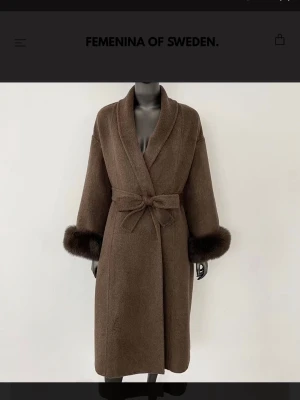 Brun kappa med pälskant från Femenina - Stilren brun kappa i längre modell från Femenina of Sweden. Kappan har bred sjalkrage, knytskärp i midjan och lyxiga pälskanter vid ärmsluten. Perfekt för dig som vill ha en elegant och trendig look.