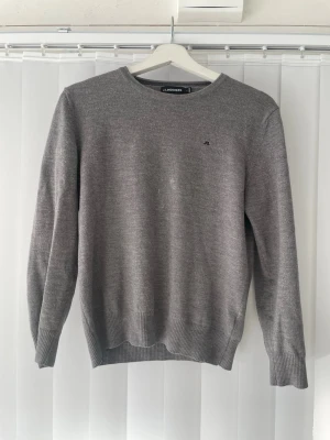 Grå tröja från J.Lindeberg - Snygg grå tröja från J.Lindeberg med rund halsringning och diskret logga på bröstet. Tröjan är stickad i mjukt material och har ribbade muddar vid ärmslut och nederkant.  Perfekt för en stilren och enkel look.  Storleken är L i barn, så passar som xs. Typ 165-170