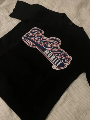 Svart BayBears Mobile t-shirt - Snygg svart t-shirt med stort BayBears Mobile-tryck i rött, vitt och blått på bröstet. Klassisk rund hals och korta ärmar. Perfekt för dig som gillar sportiga och retroinspirerade plagg.