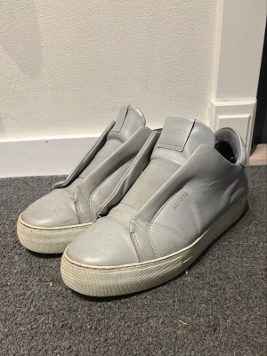 Gråa sneakers från Axel Arigato - Snygga gråa sneakers från Axel Arigato i stilren design med clean look och breda paneler. Skorna är i läder med vit sula och har diskret logga i guld på sidan. Perfekta för dig som gillar minimalistisk stil och vill ha något unikt på fötterna.