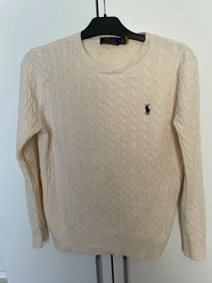 Kabelstickad beige tröja Polo Ralph Lauren - Kabelstickad beige tröja från Polo Ralph Lauren med rund halsringning och klassisk broderad logga på bröstet. Tröjan har långa ärmar och ribbade muddar. Perfekt för en stilren och avslappnad look.