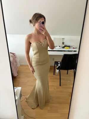 Balklänning Guldig långklänning - Säljer en elegant guldig långklänning med glittriga detaljer vid v-ringningen. Klänningen är axelbandslös och har en snygg öppen rygg med cut-out. Passformen är slim och materialet har en skimrande finish som ger en lyxig känsla. Köpt utomlands! Den är en M/L