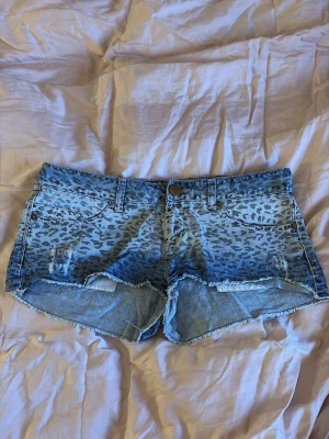 Leopardmönstrade jeansshorts - Snygga blå jeansshorts med leopardmönster och råa kanter. Shortsen har klassisk femficksdesign, bälteshällor och knappstängning framtill. Perfekta för dig som vill sticka ut med en cool och trendig look.