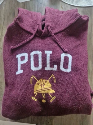  Polo Ralph Lauren hoddie  - Stickad vinröd hoodie med huva och dragsko från Polo Ralph Lauren. Stor vit POLO-text framtill och broderad gul polohjälm med klubbor. Ribbstickade muddar och nederkant. Perfekt för en avslappnad och sportig look.