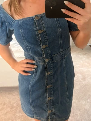 Jeansklänning med knappar - Snygg kort jeansklänning med offshoulder och tryckknappar framtill. Klänningen har markerad midja och korta ärmar. Perfekt för dig som gillar en trendig och avslappnad stil.
