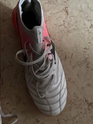 Vita och rosa Mizuno fotbollsskor - Säljer ett par snygga fotbollsskor från Mizuno i vitt och rosa med svart logga på sidan. Skorna har klassisk snörning och ovandel i syntetmaterial med detaljerade sömmar för extra känsla på bollen. Perfekta för dig som vill sticka ut på planen!
