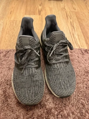Adidas grå Ultraboost sneakers - Grå Adidas Ultraboost sneakers med stickad ovandel och klassiska tre ränder i plast på sidorna. Skorna har en vit Boost-sula och grå snören. Ovandelen är i textil och sidorna i plast, vilket ger en sportig och modern look.