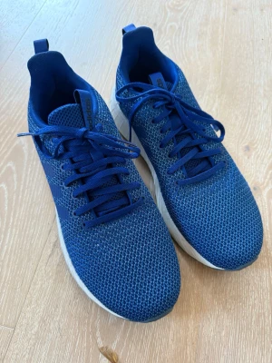 Blåa Adidas sneakers med mesh - Snygga blå sneakers från Adidas med ovandel i ventilerande mesh och klassiska tre ränder på sidan. Skorna har vita sulor och blåa skosnören. Perfekta för en sportig och stilren look. Storlek 44