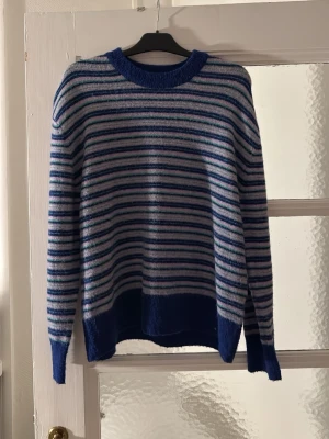 Blå- och vitrandig stickad tröja - Säljer en stickad tröja med blå, vita och gröna ränder. Tröjan har rund halsringning, långa ärmar och bredare blå kant nedtill samt vid ärmsluten. Perfekt för dig som gillar en avslappnad och färgglad stil.