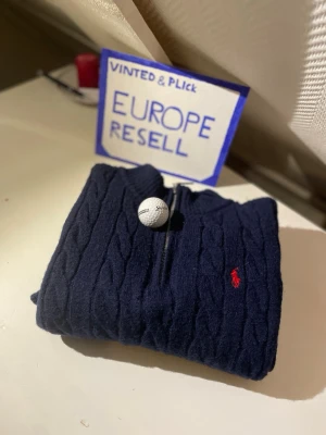 Mörkblå kabelstickad tröja Polo Ralph Lauren - Kabelstickad mörkblå tröja från Polo Ralph Lauren med halv dragkedja och röd broderad logga på bröstet. Tröjan har ribbade muddar vid ärmslut och nederkant samt hög krage. Perfekt för en klassisk och stilren look.