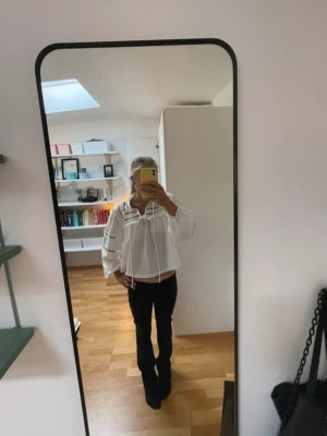 Vit sommarblus - Säljer en vit boho-inspirerad blus från H&M med långärmade puffärmar, dekorativa hålbroderier och volangdetaljer. Blusen har en lös passform och knytband vid halsen. Materialet är strukturerat och känns lätt och luftigt.