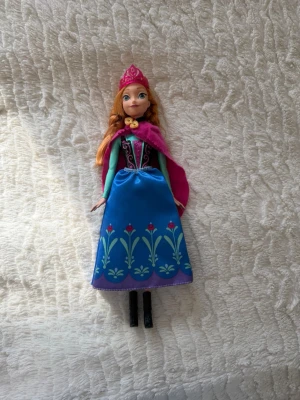 Disney Frost Anna docka med kläder och accessoarer - Disney Frost Anna docka. Dockan har rött hår flätat i två tofsar med en vit slinga i håret. Hon bär en blå klänning med blommönster, en rosa cape, en rosa krona och svarta stövlar. Kläderna är avtagbara. Dockan är märkt ”2013 Mattel” och ”Made in Indonesia”. Inget saknas dockan är komplett. I väldigt bra skick. Det ända är att håret är lite rufsigt men inte så mycket man ser på bilderna!!