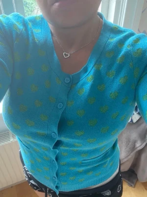 Blå kofta med gröna hjärtan - Supersöt blå kofta  med gröna hjärtan över hela plagget. Koftan har rund halsringning, långa ärmar, knäppning framtill och två små fickor. Perfekt för dig som gillar färgglada och lekfulla plagg!