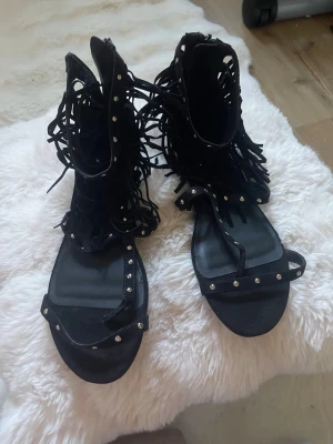 Svarta sandaler med fransar och nitar - Coola svarta sandaler med högt skaft, dekorerade med fransar och små silvriga nitar. Sandalerna har öppen tå och remmar framtill. Perfekta för dig som vill sticka ut med en edgy och trendig stil. Materialet ser ut att vara konstläder.