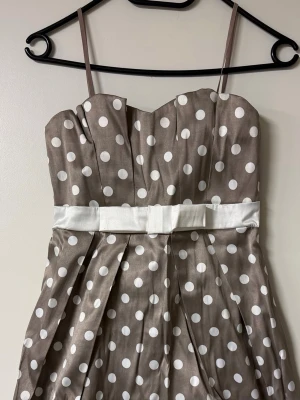 Beige prickig kortklänning med rosett - Söt beige kortklänning med vita prickar och tunna axelband. Klänningen har en markerad midja med vit rosett och veckad kjol för extra volym. Perfekt för dig som gillar retrovibbar och lekfull stil.